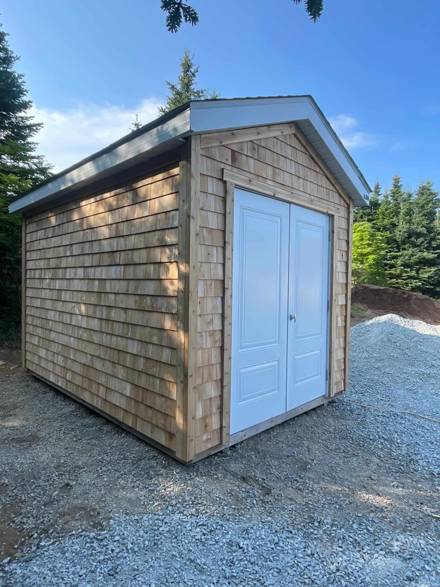 A cedar-shingled shed seen from the corner, with a gable roof, white fascia and a pair of white double doors, standing on a gravel pad among spruce trees.