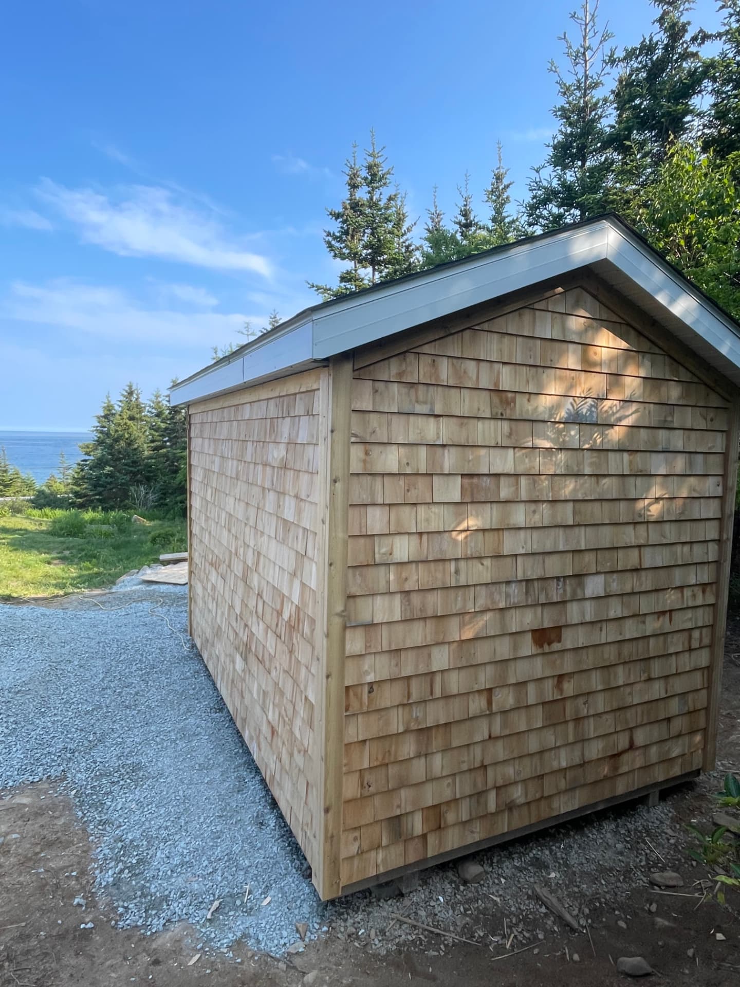 The rear gable end of a cedar-shingled shed, coursed shingles running up to the peak, with the ocean visible past the trees.