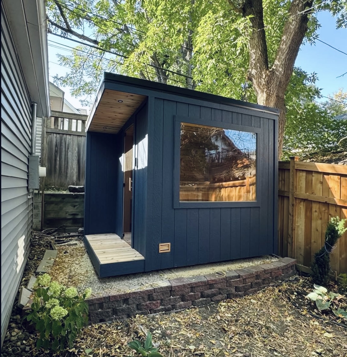 A compact backyard sauna with dark navy vertical board siding, a large fixed picture window, cedar soffit and a cedar entry step.