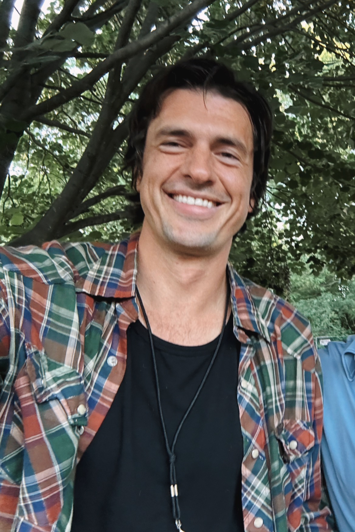 Drake Ritter smiling in a plaid shirt under a leafy tree.