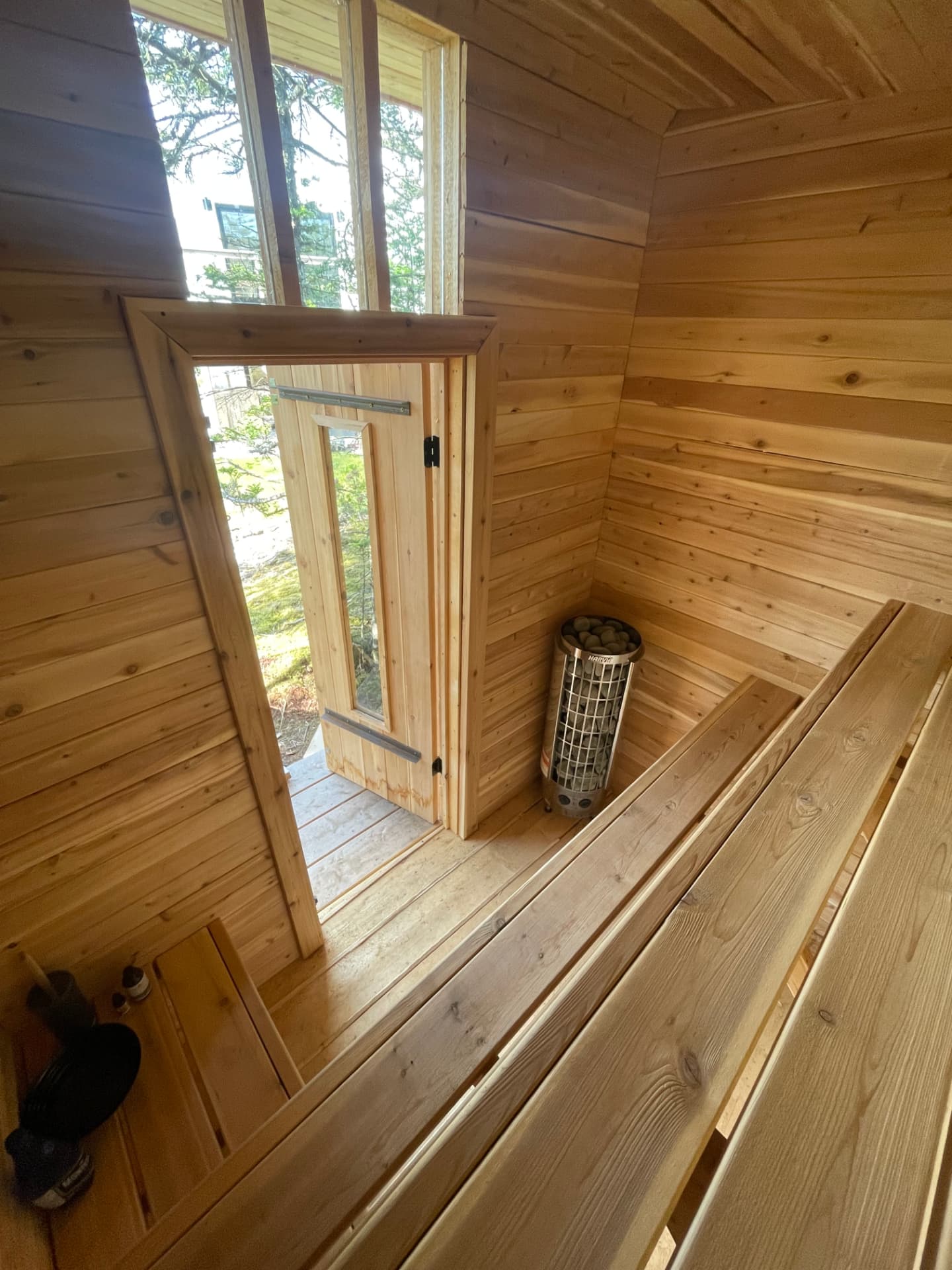 The interior of a cedar sauna looking down at an open plank door with a glass panel, a tall window above, a stone-filled heater in the corner and two tiers of cedar benches.