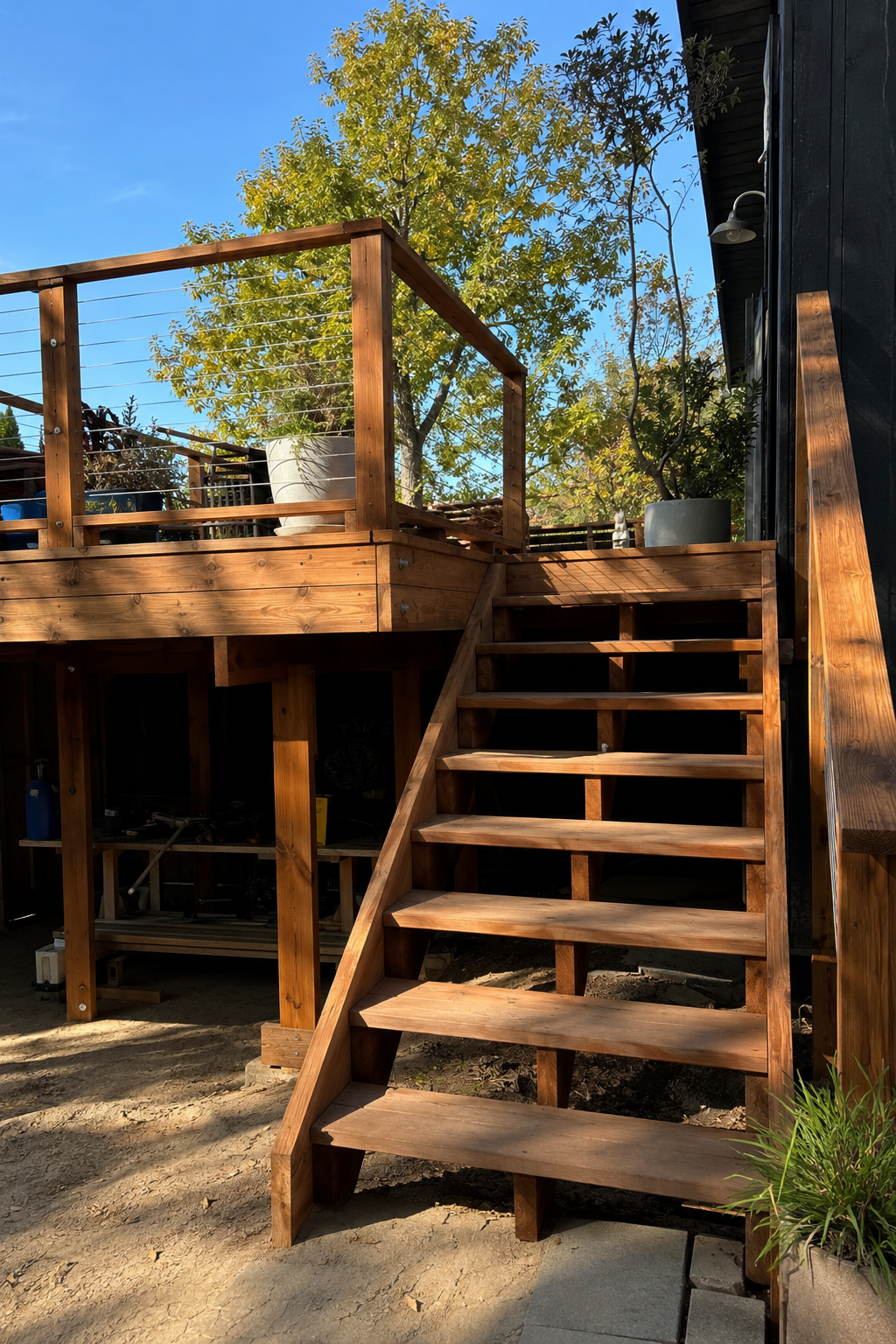 A redwood stair run with open risers climbing to an elevated deck with cable railing, beside a black-clad house.