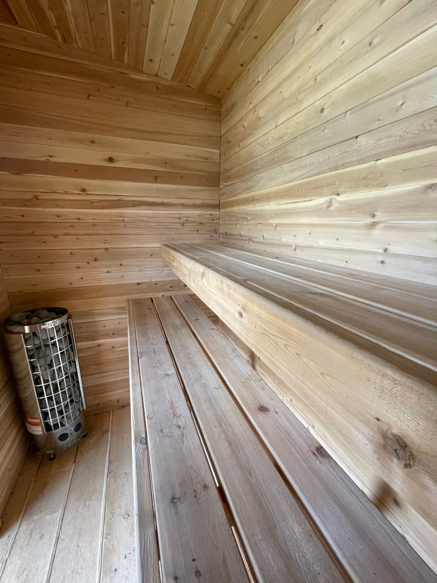 Two tiers of cedar benches inside a sauna clad in horizontal boards, with a stone-filled heater at the left.