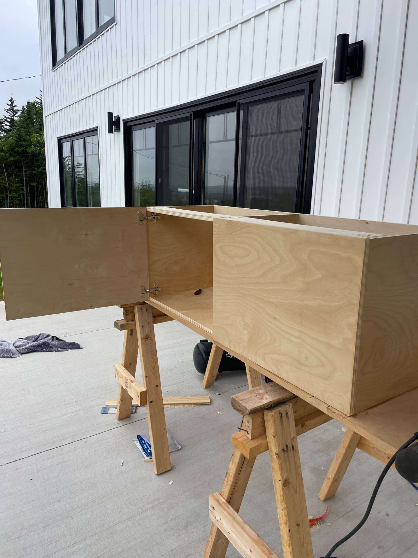 A plywood cabinet carcass with a hinged door standing on two wooden sawhorses outside a white building.