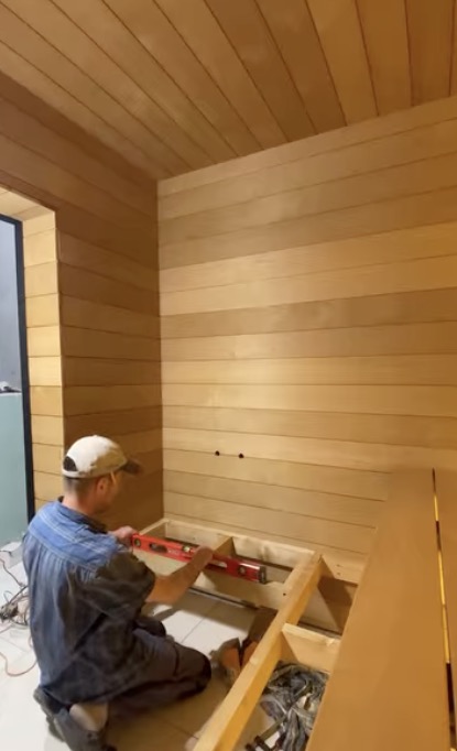 A carpenter checking a level on a bench frame inside a sauna room clad in horizontal light-toned boards.