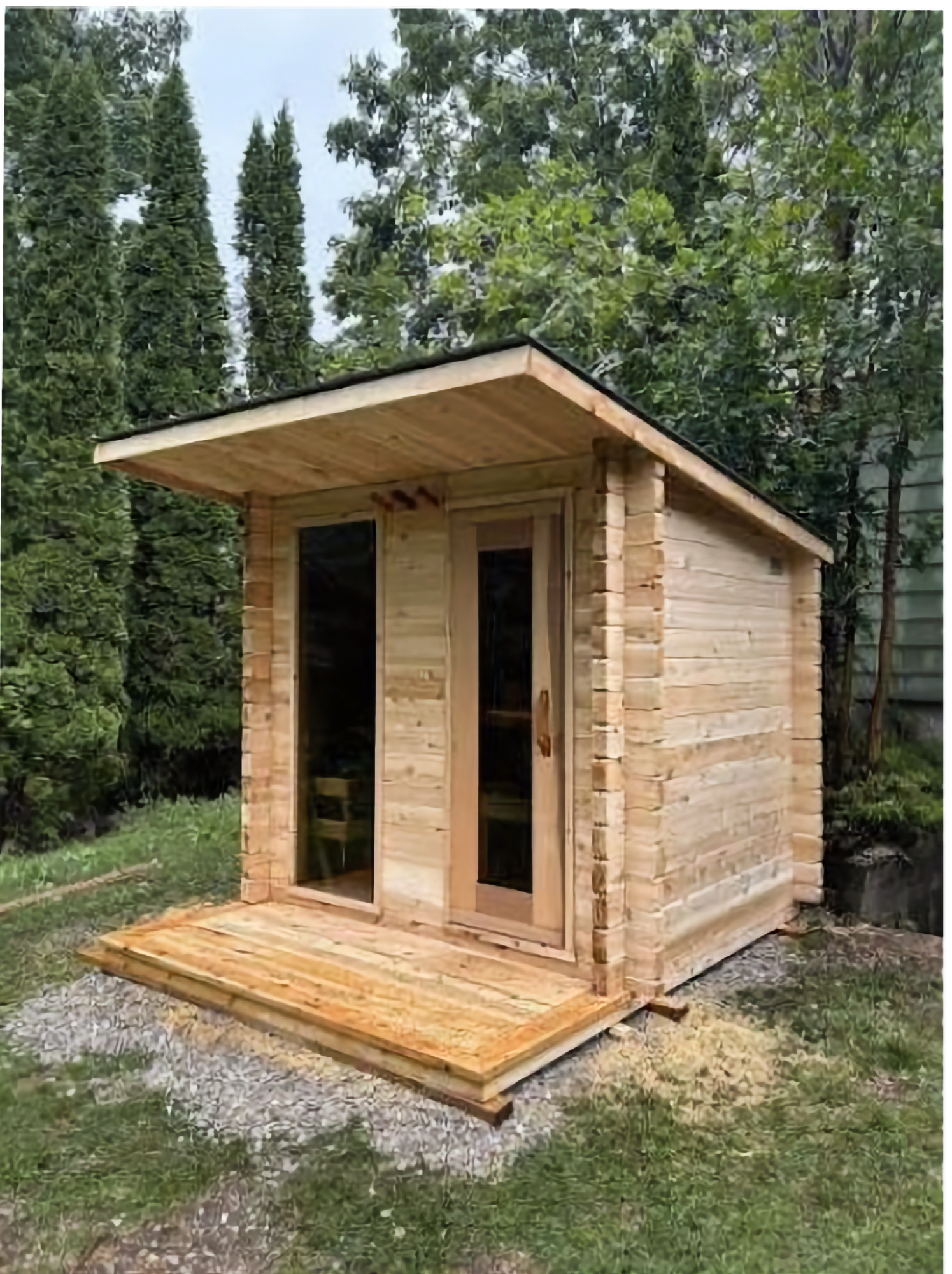 A small cedar outbuilding with log-profile siding, a glass door and side window, a deep roof overhang and a cedar deck at the entry.
