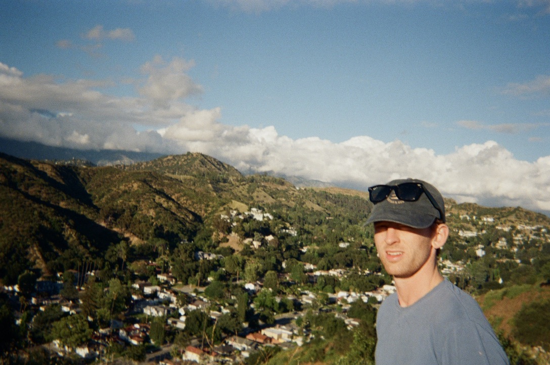 Ian Cazador in a cap and sunglasses on a ridge above a hillside Los Angeles neighborhood.