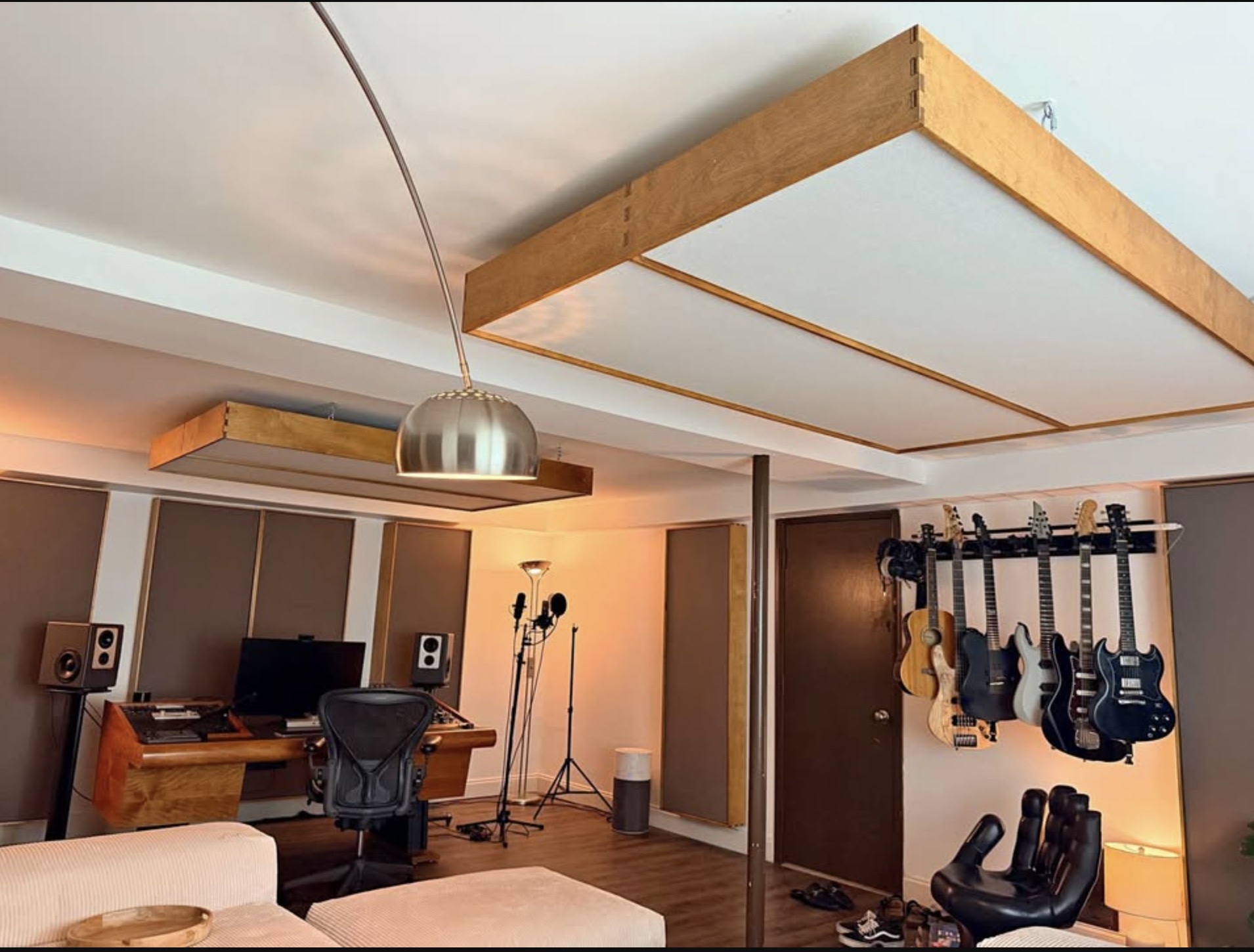 Large birch-framed acoustic ceiling clouds suspended over a home studio mix desk, with wall panels, monitors and guitars hung on the far wall.